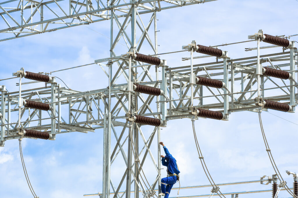Electrician is climbing on high voltage electric pylon structure to installing electrical equipment in power distribution substation against blue sky background